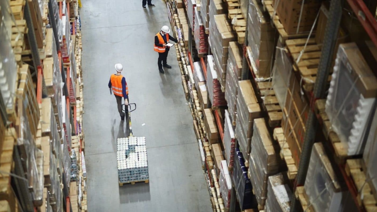 Aerial view of two workers in safety vests and helmets in a warehouse aisle, one pulling a pallet jack with boxes, the other holding a clipboard among tall shelves filled with neatly stacked packages.
