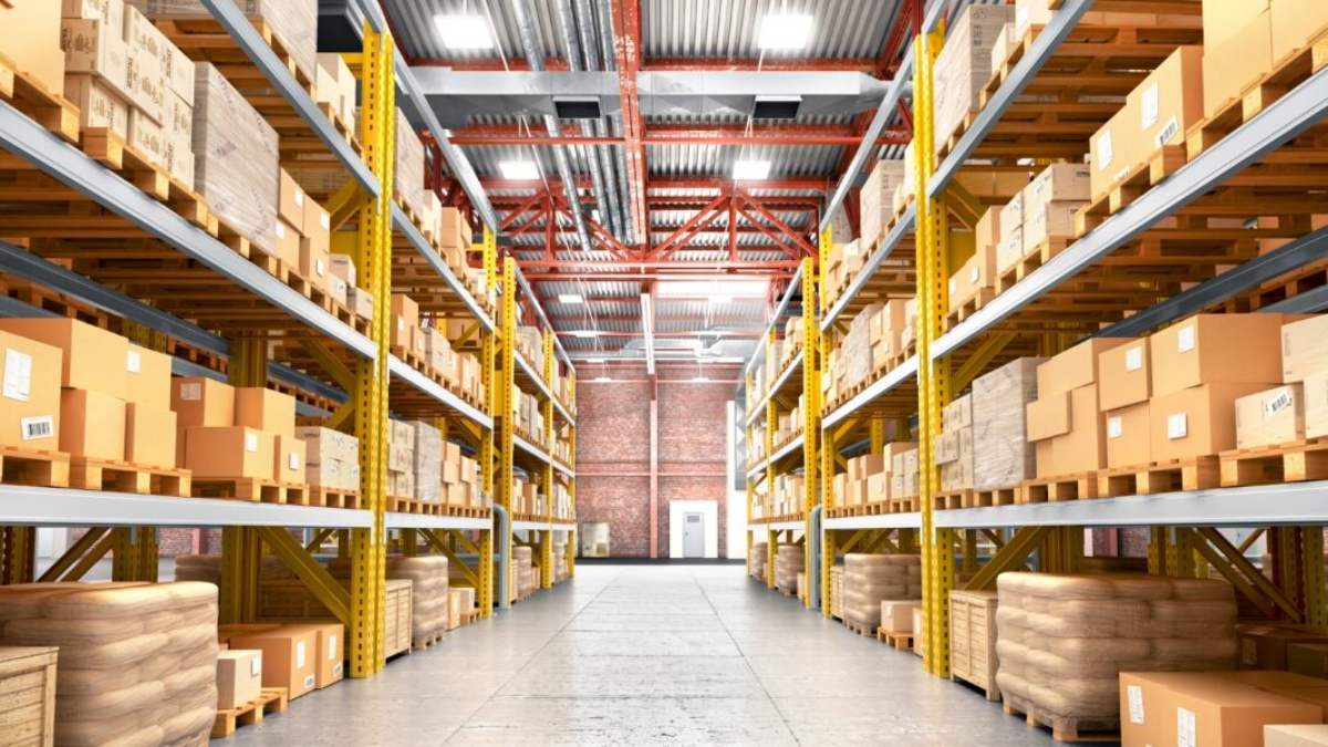 A spacious warehouse with tall shelves filled with cardboard boxes and packages. Robotics integration streamlines operations, while organized rows and a clear concrete floor create an orderly environment brightly illuminated throughout.