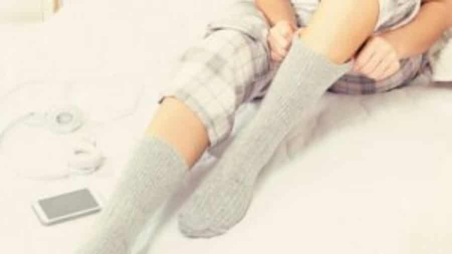 A person wearing plaid pajamas sits on a bed, pulling up gray socks. A smartphone and white headphones lie beside them.