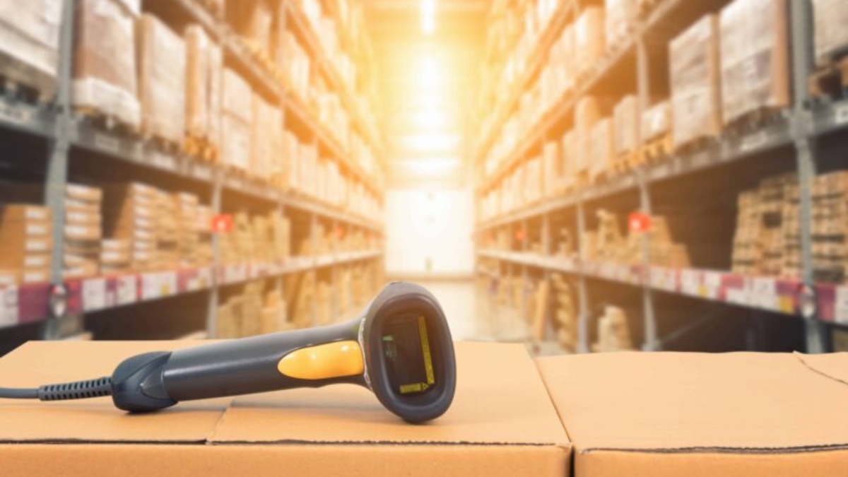 A barcode scanner resting on stacked cardboard boxes in a warehouse, surrounded by shelves filled with various boxes in the background, highlights efficient inventory management under bright sunlight.
