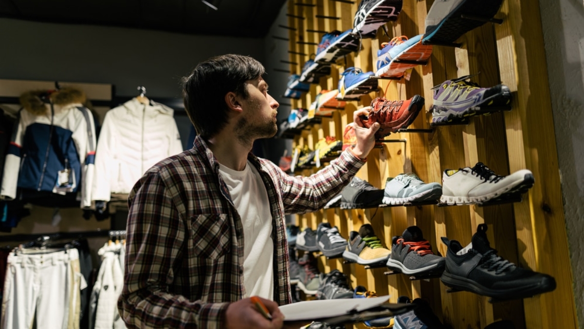 A man in a plaid shirt holds a clipboard and examines running shoes displayed on a wooden wall, focused on assortment planning in the sports store. Clothing and jackets hang neatly on racks in the background.
