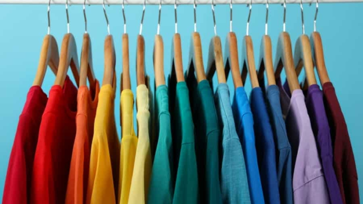 A row of colorful t-shirts hanging on wooden hangers, arranged in a gradient rainbow order, against a light blue background.