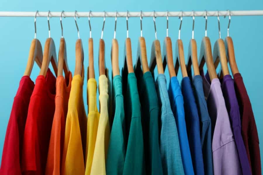 A row of colorful t-shirts hanging on wooden hangers, arranged in a gradient rainbow order, against a light blue background.