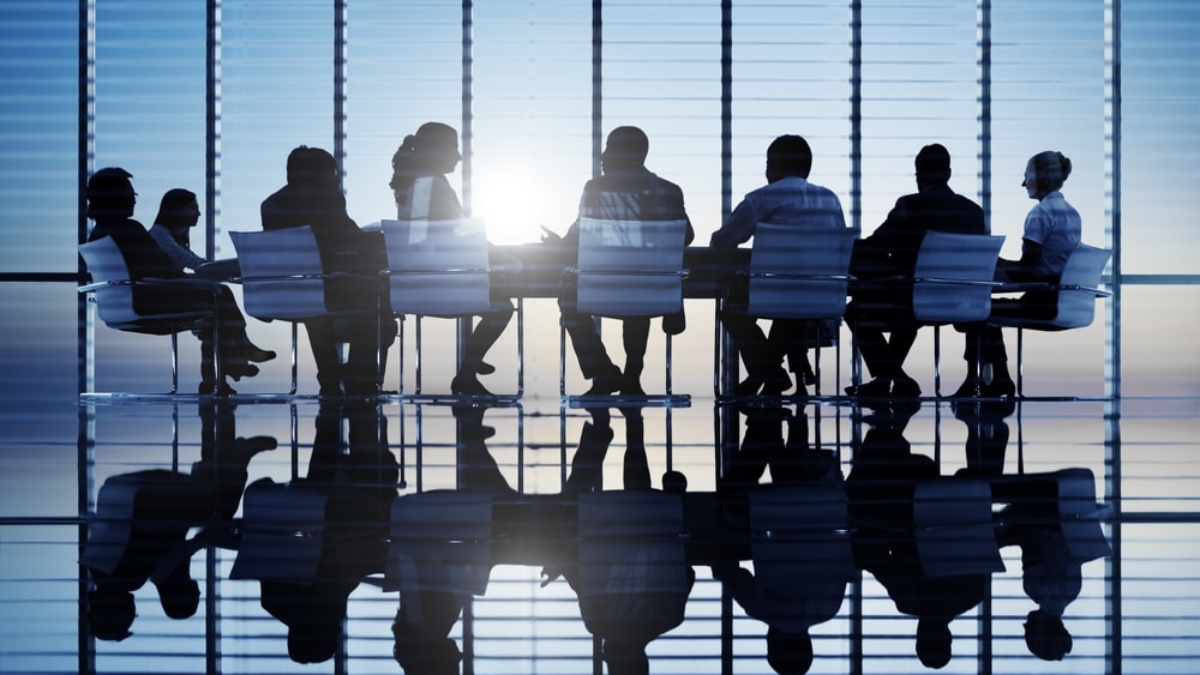 A group of people in business attire sit around a conference table in a modern office with large windows, silhouetted against the sunlight, with their reflections visible on the shiny floor.
