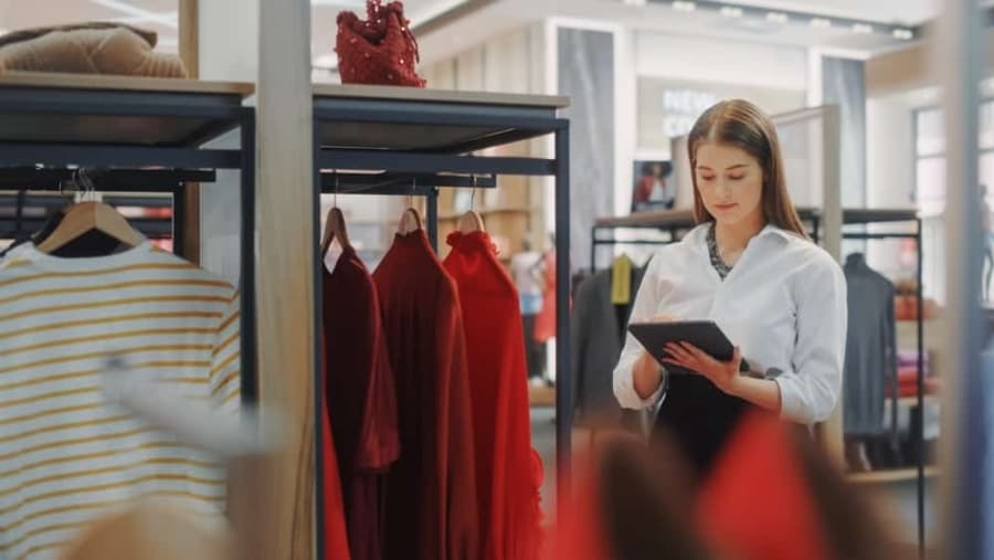 A woman in a white blouse stands in a clothing store, holding and using a tablet. She is surrounded by racks of clothes, including red dresses and a striped shirt, and appears focused on her task.