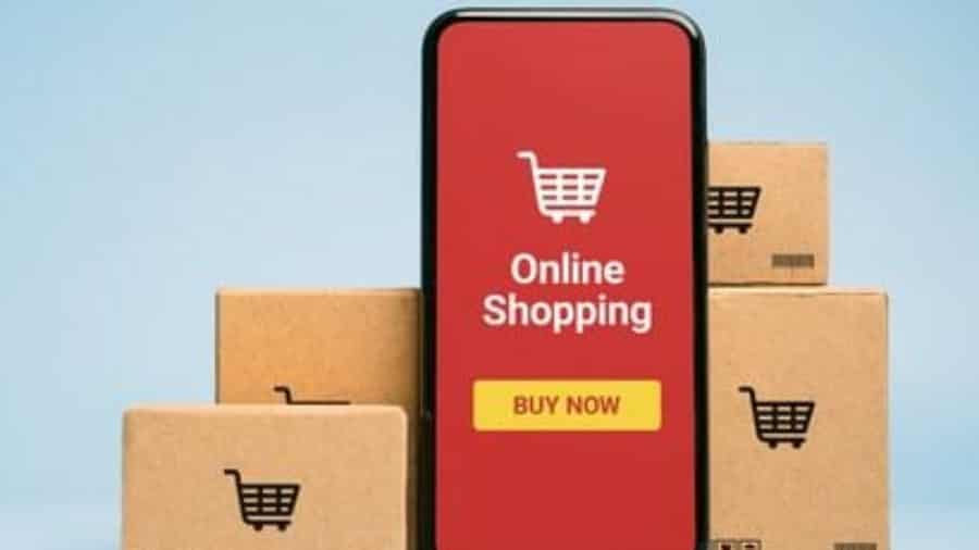 A smartphone displaying a red screen with a shopping cart icon, the words Online Shopping, and a Buy Now button, surrounded by several cardboard boxes with shopping cart symbols.