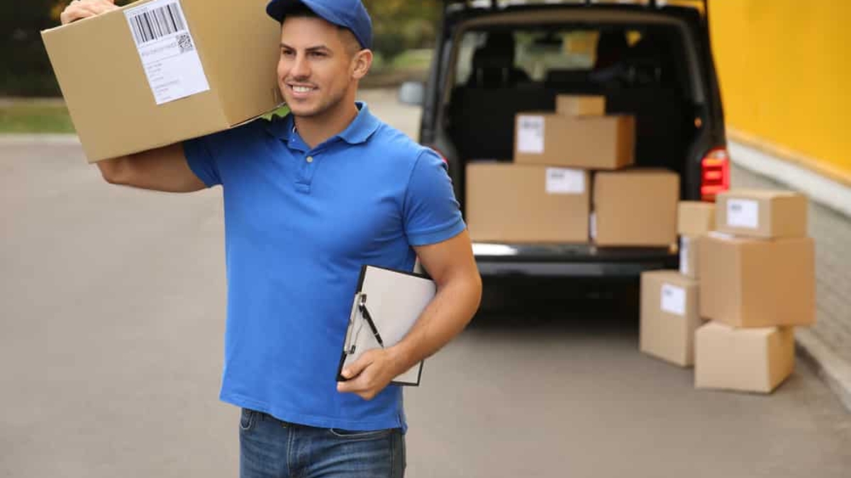 A delivery person in a blue uniform carries a large cardboard box on his shoulder and holds a clipboard, standing near a van with several packages loaded and stacked on the ground behind him.