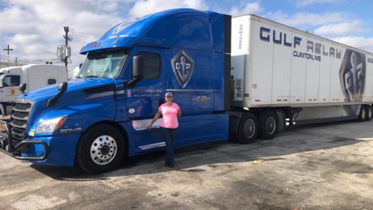 A woman in a pink shirt and cap stands beside a large blue semi-truck with Gulf Relay Clinton, MS written on the white trailer, parked in a lot under a partly cloudy sky.