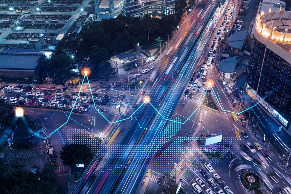 Aerial view of a busy city intersection at night with light trails from moving vehicles and digital data graphics, including a blue line graph and data points, overlaying the image to suggest smart city technology or traffic analytics.