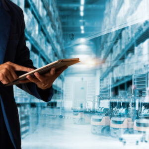 A person in a blue suit uses a tablet, with a modern warehouse filled with shelves and forklifts in the background, symbolizing logistics, inventory management, or supply chain planning operations.