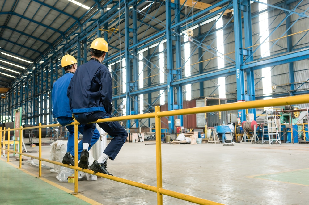Two workers in blue uniforms and yellow hard hats sit on a yellow metal railing inside a spacious industrial warehouse with high ceilings and blue structural beams.