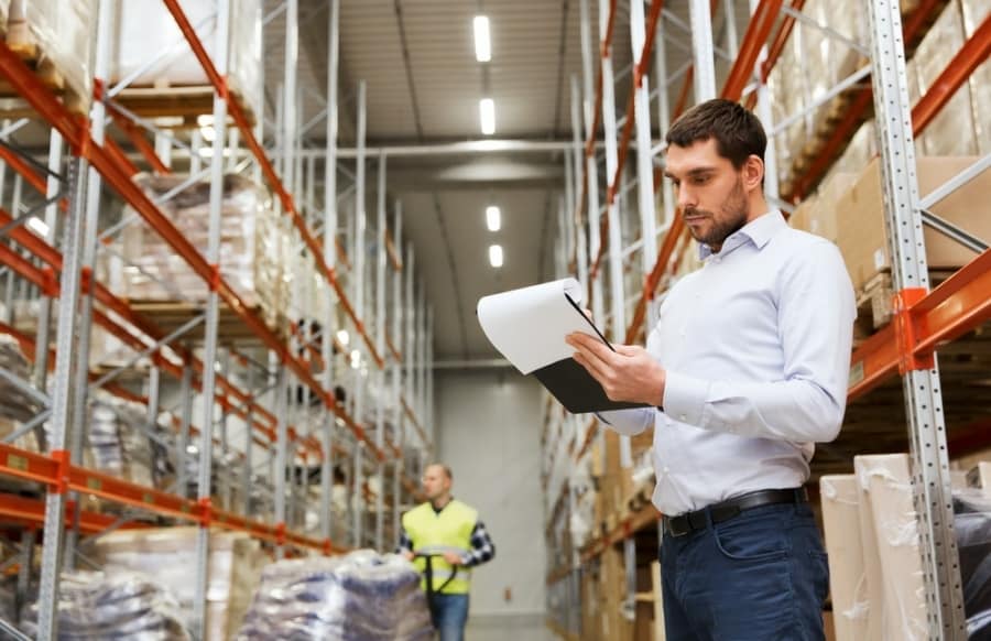 A man in a white shirt holds a clipboard and checks inventory in a large warehouse with tall shelves, while another worker in a safety vest walks in the background.