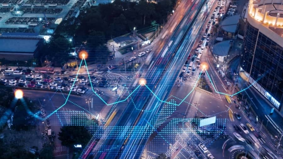 A busy city intersection at night with light trails from cars. Digital blue and orange lines and data points overlay the scene, representing data analytics or smart city technology tracking traffic.