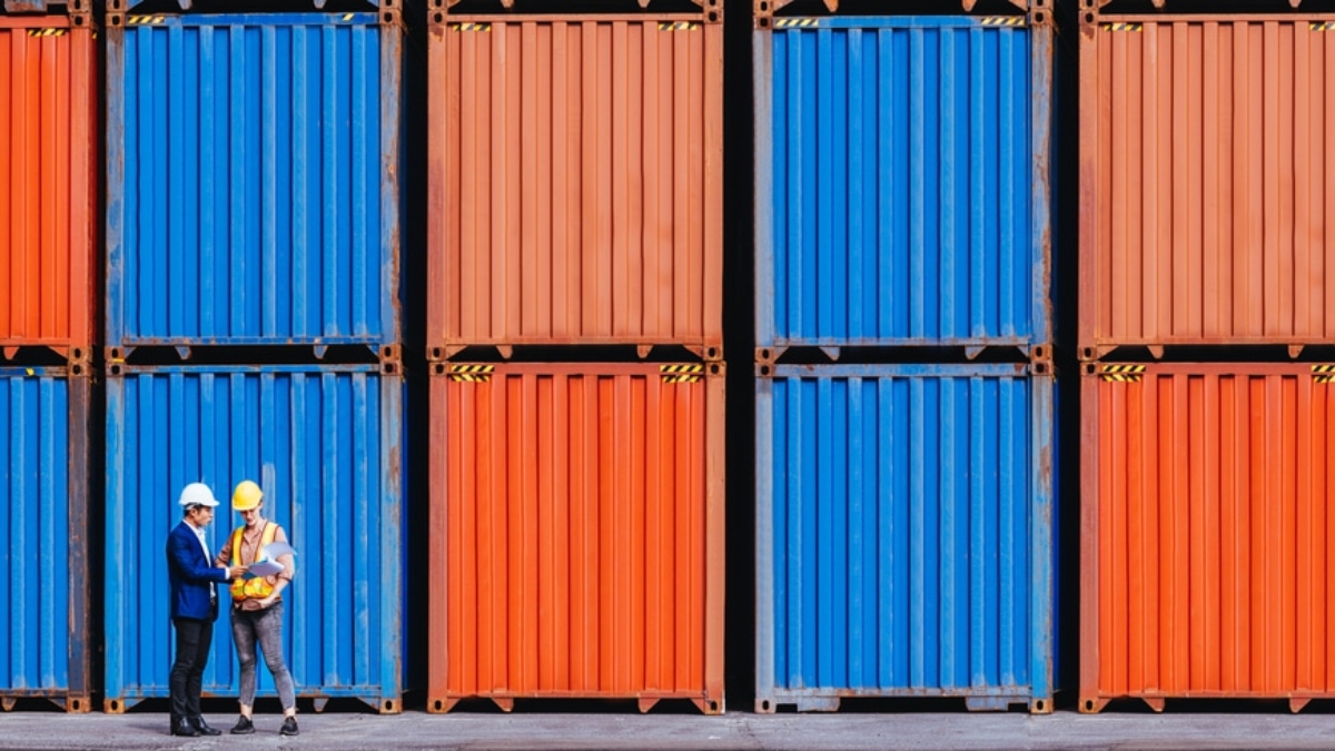 Two people wearing helmets stand in front of stacked orange and blue shipping containers, appearing to have a discussion at a shipping yard or port.
