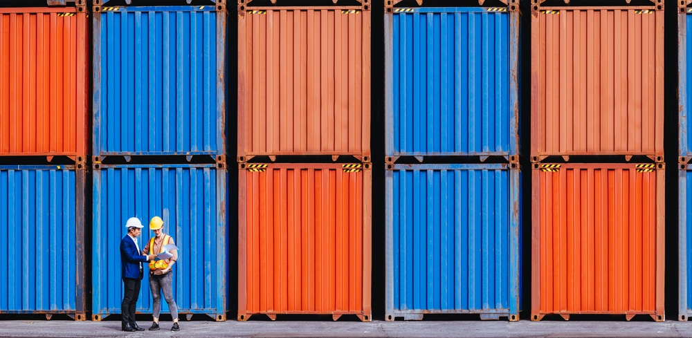 Panoramic,Of,Smart,Creative,Foreman,And,Engineer,Woman,Control,Loading Two people wearing helmets stand in front of stacked orange and blue shipping containers, appearing to have a discussion at a shipping yard or port.