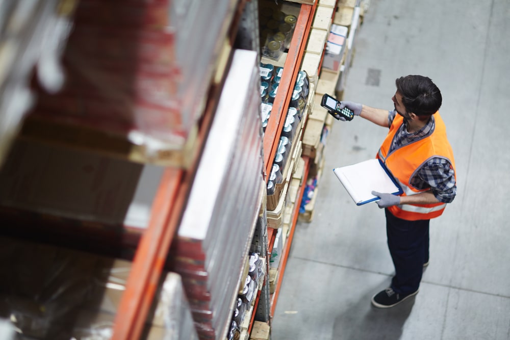 A warehouse worker in an orange safety vest scans items on a high shelf with a handheld device while holding a clipboard, standing in an industrial storage area.