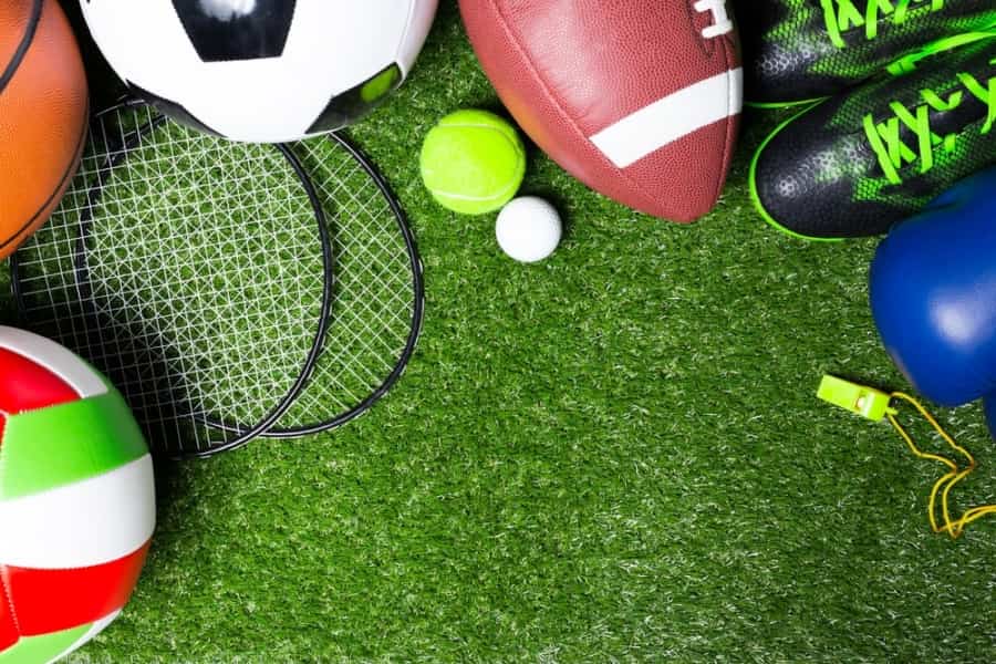 Various sports equipment, including soccer ball, football, volleyball, tennis rackets, tennis ball, golf ball, green sports shoes, blue boxing glove, and a yellow whistle, arranged on green artificial grass.
