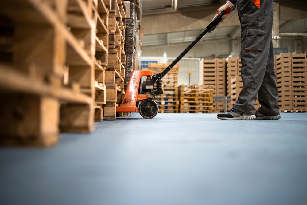 Working,At,Warehouse.,Low,Angle,View,Of,Unrecognizable,Worker,Lifting A worker in gray pants and sneakers operates an orange pallet jack, moving wooden pallets inside a warehouse with stacks of pallets in the background.