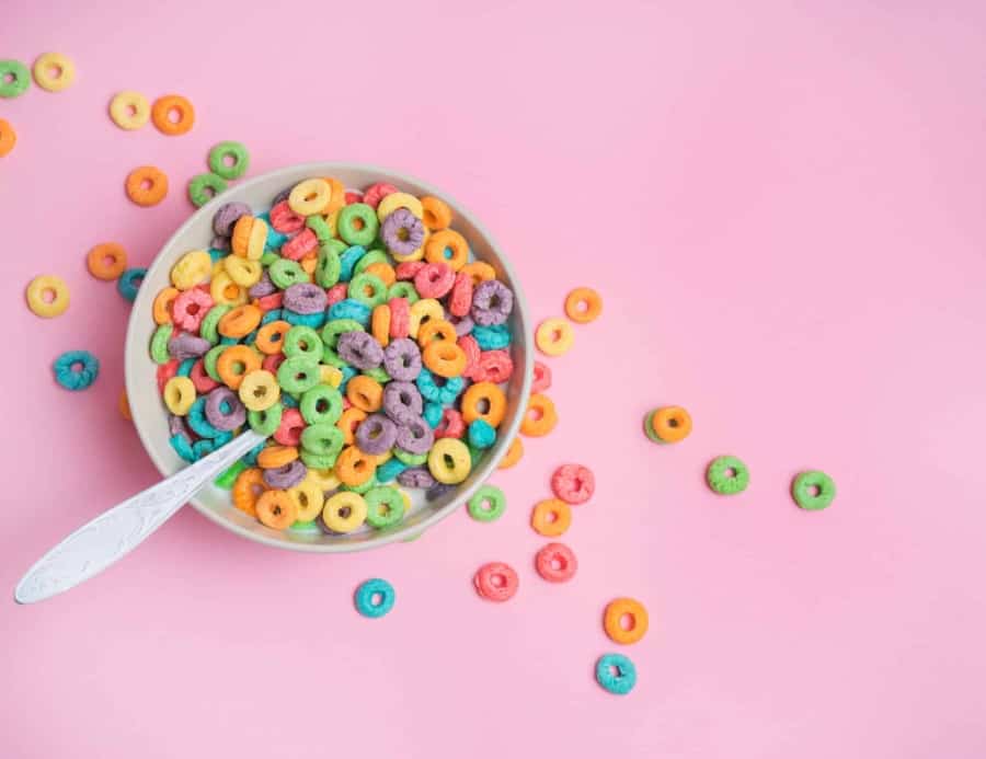 A bowl of colorful, round fruit-flavored cereal with a spoon sits on a pink background, with several cereal pieces scattered around the bowl.