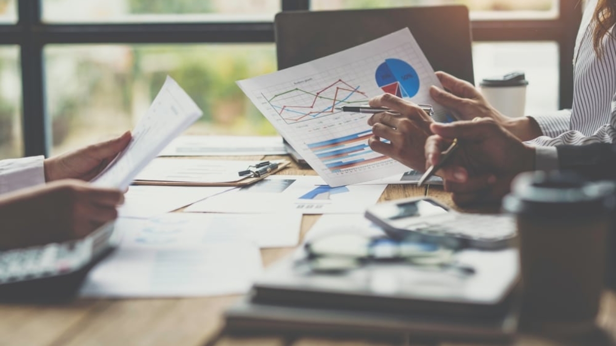Three people sit at a table reviewing documents and charts with graphs and data. Laptops, coffee cups, and office supplies are on the table, suggesting a business meeting or team discussion in a bright office setting.
