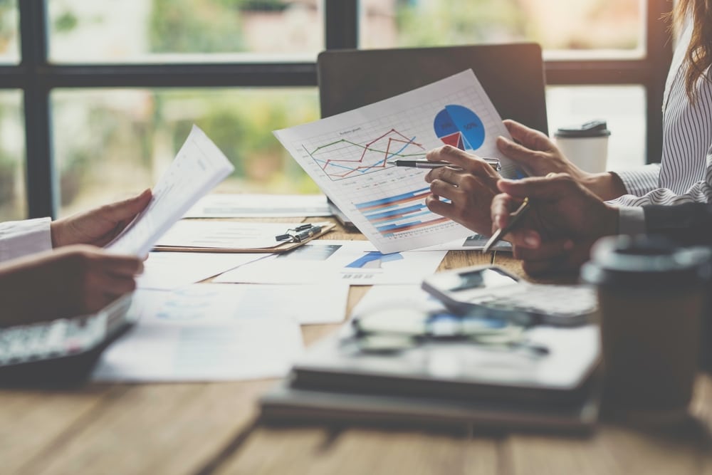 Three people sit at a table reviewing documents and charts with graphs and data. Laptops, coffee cups, and office supplies are on the table, suggesting a business meeting or team discussion in a bright office setting.
