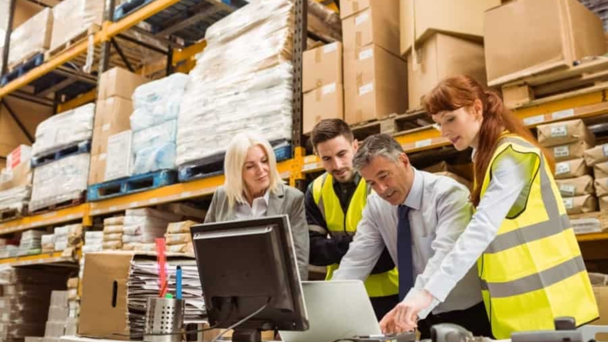 Four warehouse workers, three wearing yellow safety vests, gather around a desk with a computer and paperwork, discussing something in a busy storage facility with shelves stacked high with boxes and packages.