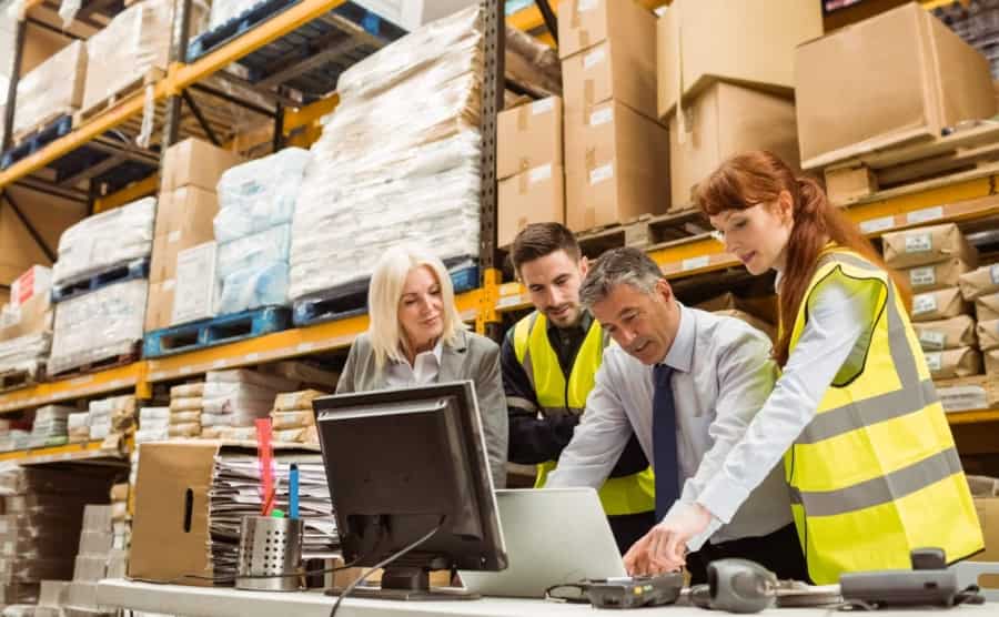 Four warehouse workers, three wearing yellow safety vests, gather around a desk with a computer and paperwork, discussing something in a busy storage facility with shelves stacked high with boxes and packages.