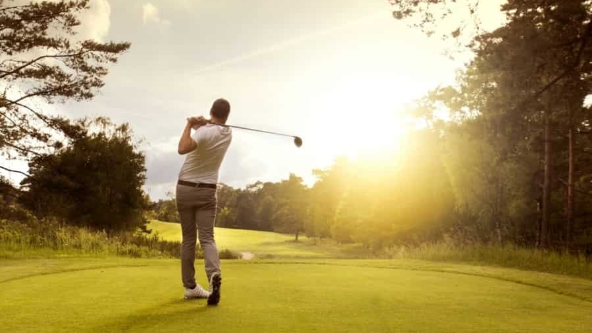 A person swings a golf club on a grassy course at sunset, surrounded by trees and a bright sky.