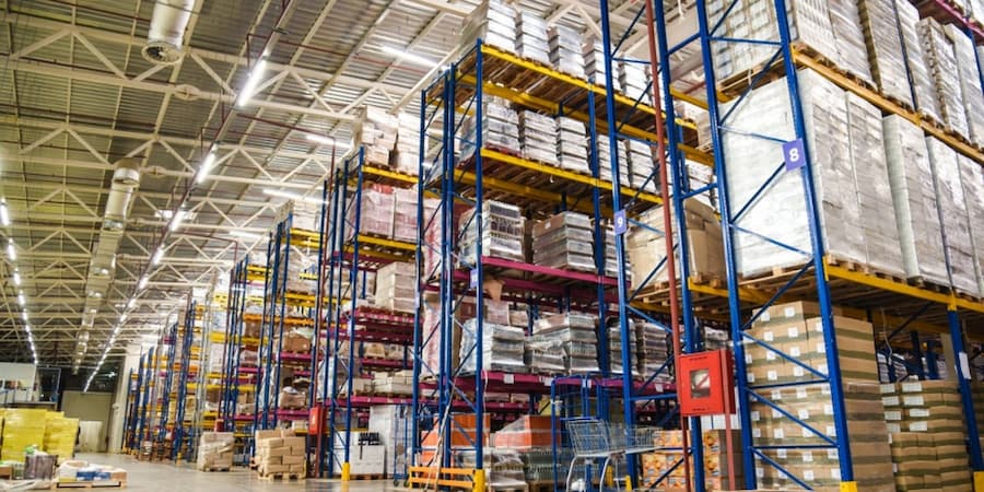 A large warehouse with tall shelves stacked with pallets of goods, boxes, and packages. The shelves are color-coded in blue and yellow. The warehouse is brightly lit with overhead lighting.