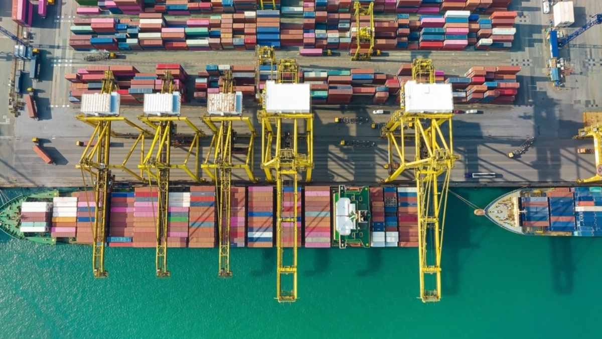 Aerial view of a busy shipping port with yellow cranes loading and unloading colorful cargo containers onto docked container ships along a turquoise waterfront.