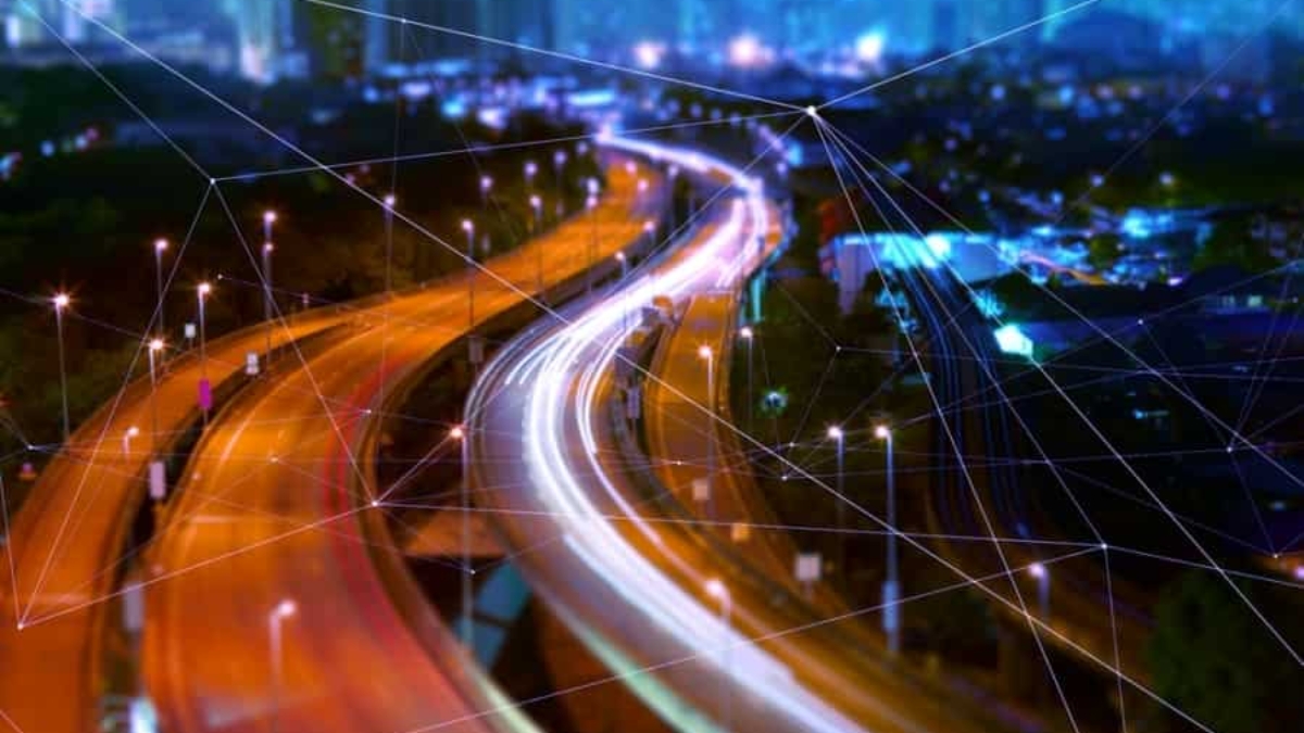 A city highway at night with bright, colorful light trails from moving vehicles. Skyscrapers rise in the background as translucent digital lines and nodes overlay the scene, hinting at a connected future powered by a Transportation Management System.