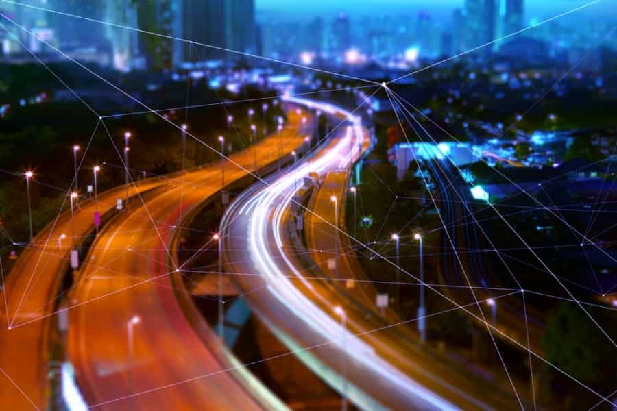 A city highway at night with bright, colorful light trails from moving vehicles. Skyscrapers rise in the background as translucent digital lines and nodes overlay the scene, hinting at a connected future powered by a Transportation Management System.