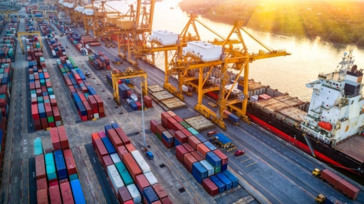 Aerial view of a busy 3PL shipping port with large yellow cranes, stacked colorful shipping containers, and a cargo ship docked by the water at sunset.