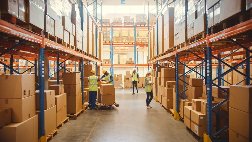 shutterstock_1848611221-6 (1) Warehouse workers in safety vests organize and move large cardboard boxes along wide aisles lined with tall metal shelves, efficiently tracking inventory with a WMS. Bright lighting and high ceilings are visible.