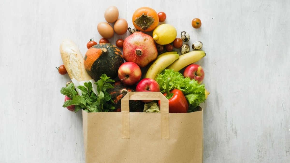 A brown paper bag overflowing with fresh groceries, including eggs, bread, pumpkins, apples, bananas, lettuce, tomatoes, a pomegranate, lemons, strawberries, mint, and quail eggs on a white surface—perfect for FSMA-compliant kitchens.