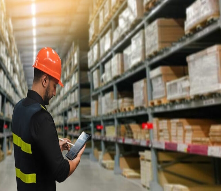 A warehouse worker wearing a safety vest and orange hard hat uses a tablet while standing in an aisle lined with tall shelves stocked with various materials.