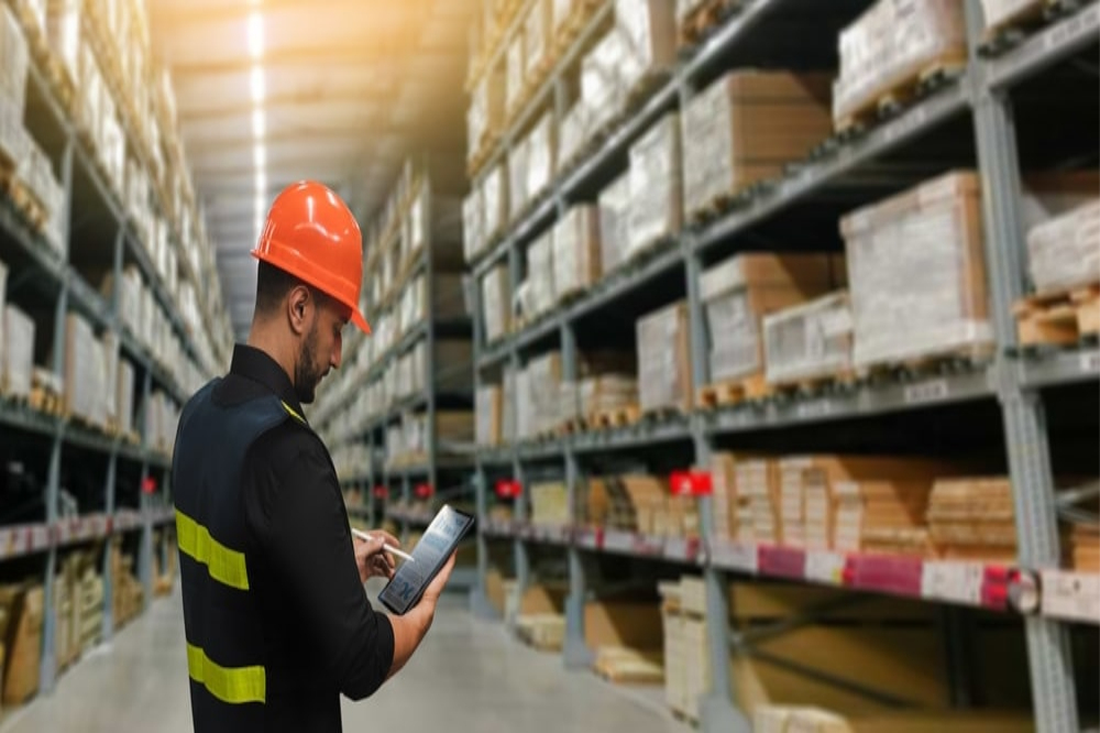 A warehouse worker wearing a safety vest and orange hard hat uses a tablet while standing in an aisle lined with tall shelves stocked with various materials.