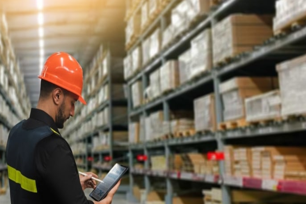A worker wearing an orange hard hat and safety vest uses a tablet in a warehouse filled with shelves stacked with boxes and packages.