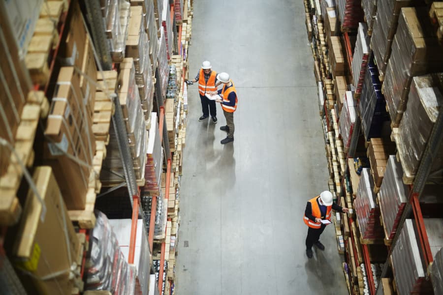 Overhead view of three workers in orange safety vests and hard hats walking and inspecting shelves in a large warehouse filled with stacked boxes and packages.