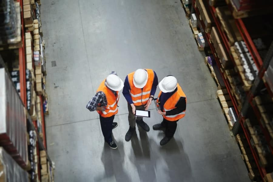 Three people wearing white hard hats and orange safety vests stand in a warehouse aisle, looking at a tablet. Shelves stocked with boxes and items are visible on both sides.