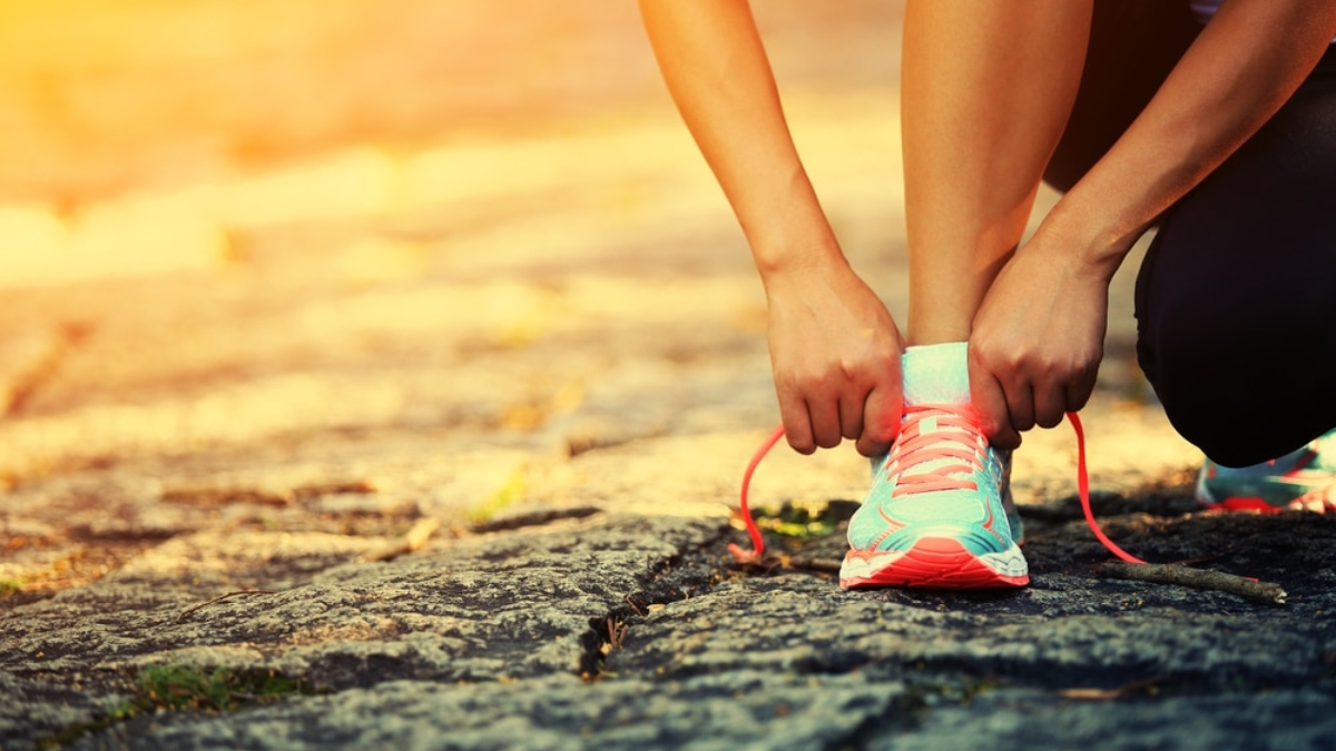 A person tying the laces of a bright teal and pink running shoe while crouching on a sunlit stone path, preparing for a run or workout.