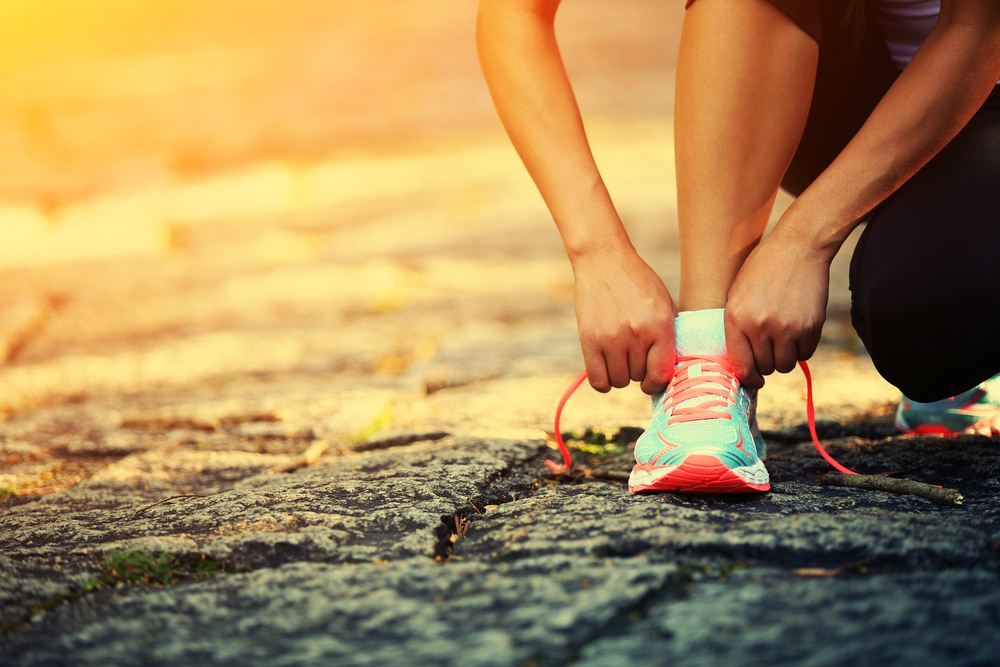 A person tying the laces of a bright teal and pink running shoe while crouching on a sunlit stone path, preparing for a run or workout.
