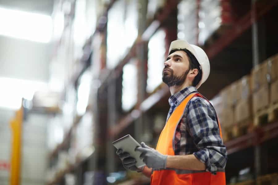 A warehouse worker in a hard hat, orange safety vest, and gloves holds a tablet, possibly checking a Warehouse Management System (WMS) Cheat Sheet while looking up at shelves full of inventory in a blurred industrial setting.