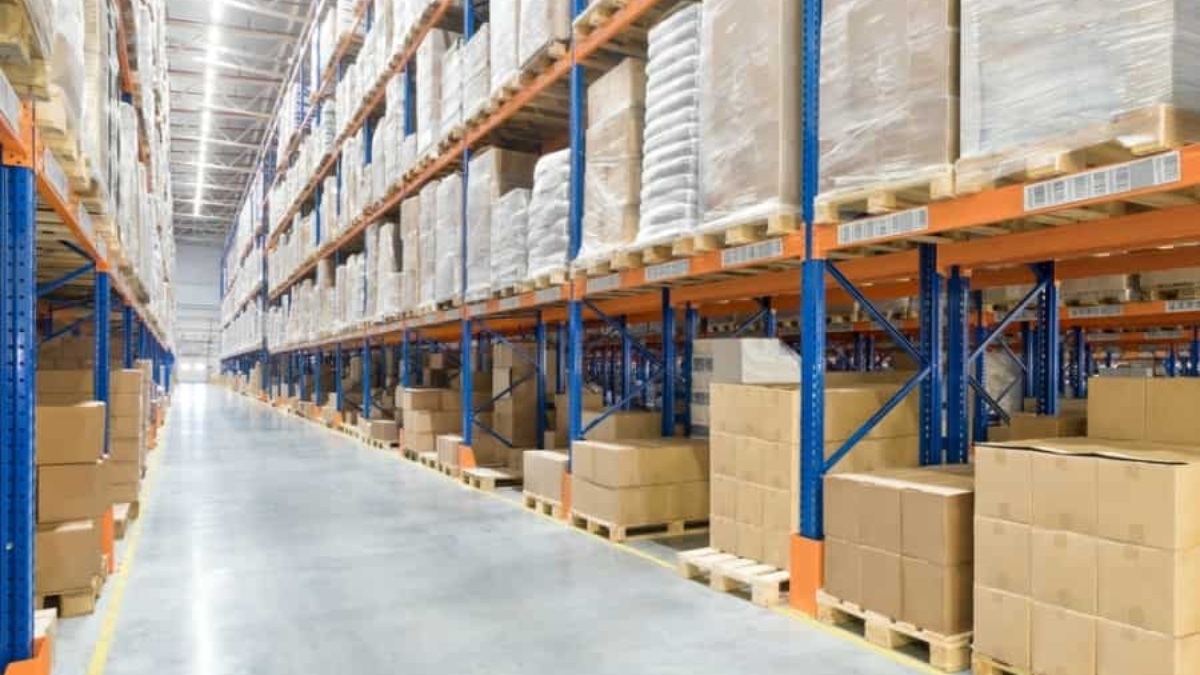 Large warehouse interior with tall blue and orange industrial shelving, stacked with cardboard boxes and wrapped pallets; the polished concrete floor and wide aisle highlight a space designed for efficient warehouse automation.