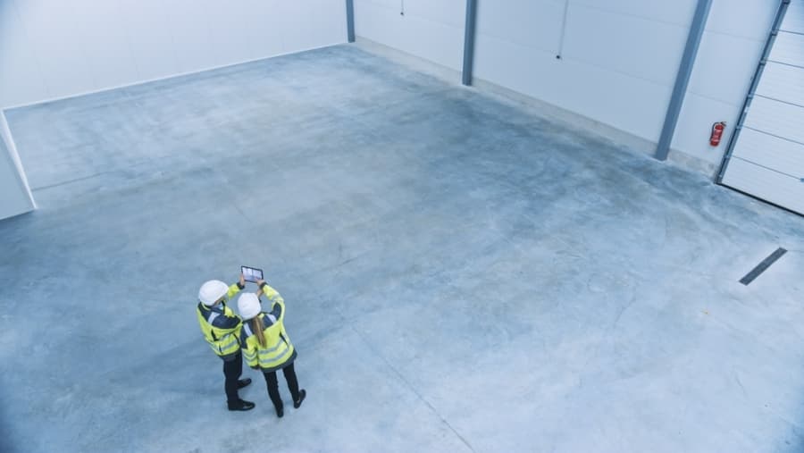 Two people in safety vests and helmets stand together in a large, empty industrial warehouse, looking at a tablet. The warehouse has concrete floors and high white walls.