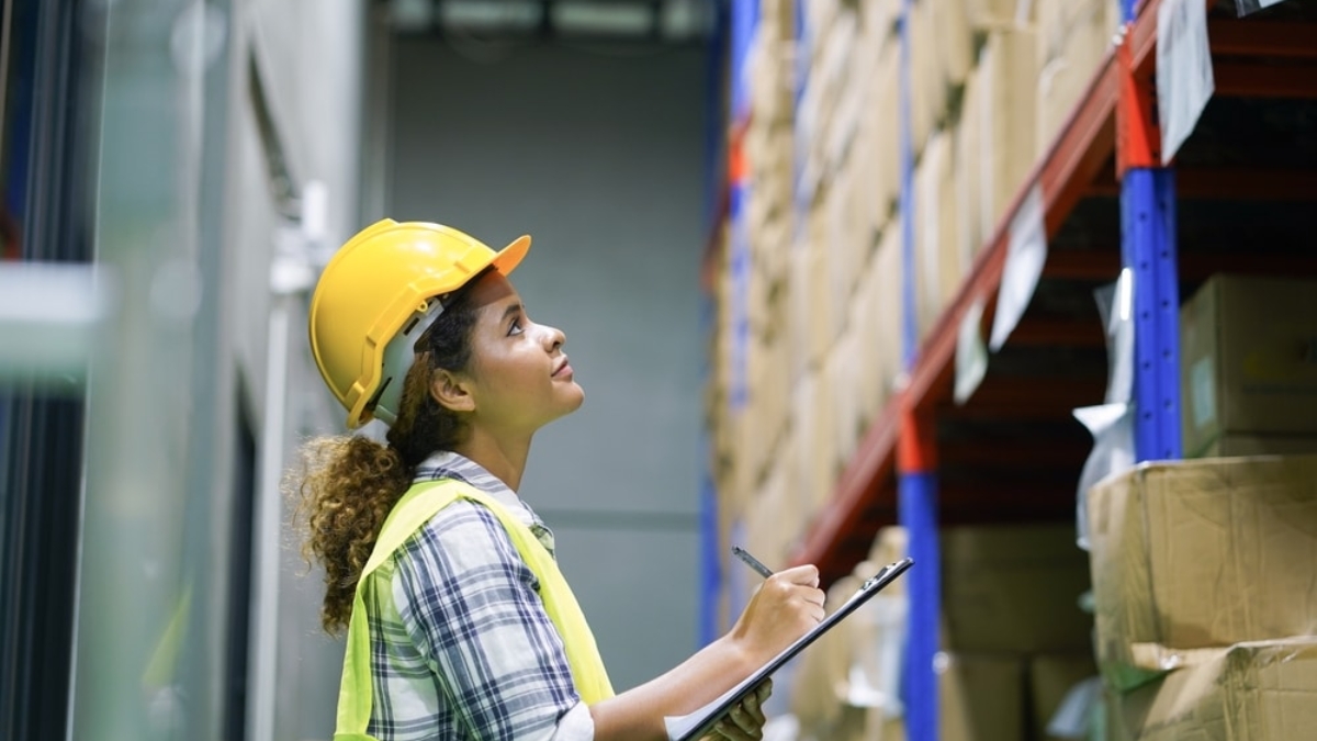 A woman wearing a yellow hard hat and safety vest stands in a warehouse, promoting warehouse safety as she holds a clipboard and pen, looking up at shelves stacked with cardboard boxes.