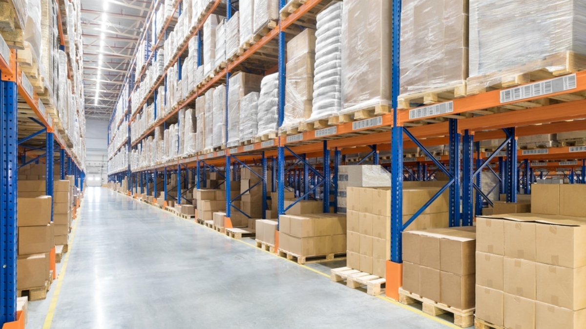 Rows of tall warehouse shelves filled with stacked cardboard boxes and pallets line a wide, clean aisle in a brightly lit industrial storage facility.