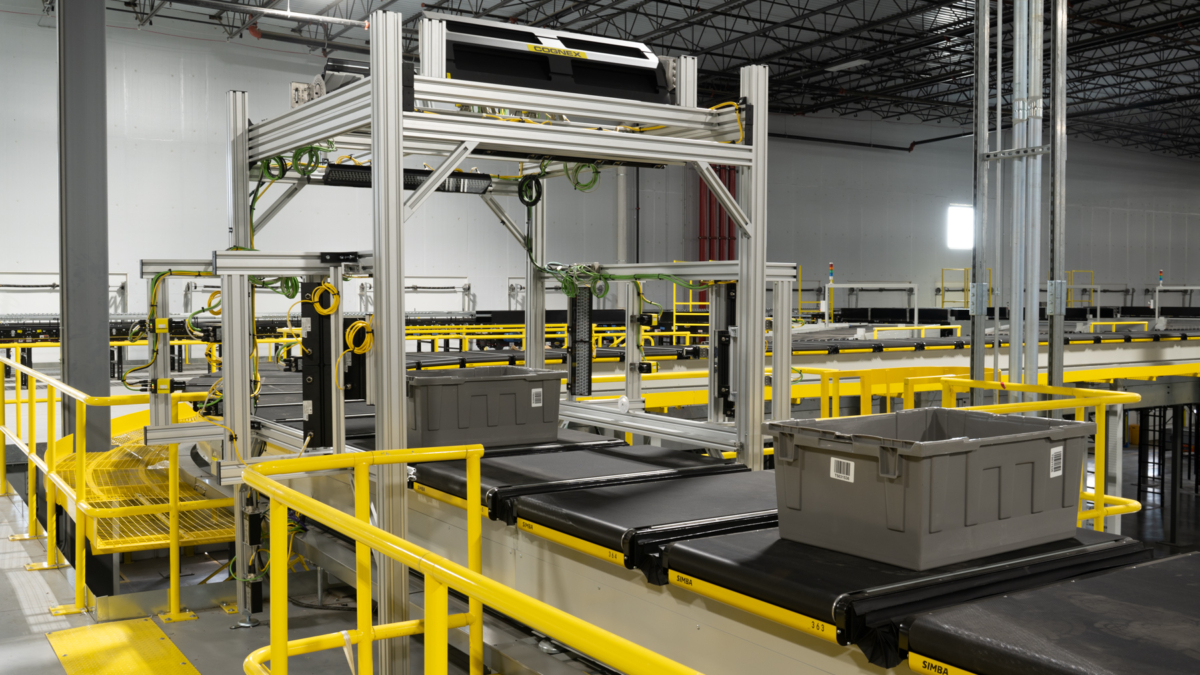 A gray plastic bin moves along a conveyor belt system inside a large industrial warehouse with yellow safety railings and exposed ceiling beams.