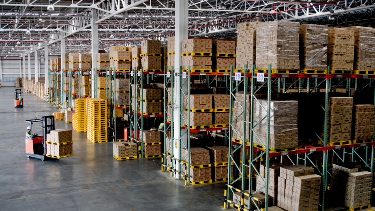 A large warehouse interior with tall shelves stacked with boxes and pallets, featuring efficient slotting and a worker driving a forklift along the aisle. The space is well-lit and organized, with industrial shelving and high ceilings.