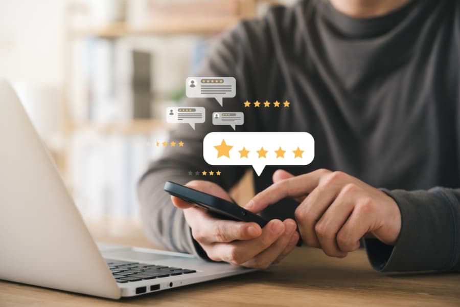 A person sits at a desk using a smartphone next to a laptop, with illustrated five-star rating and review icons floating above, suggesting online feedback or customer review activity powered by Microsoft Dynamics 365 Customer Experience.